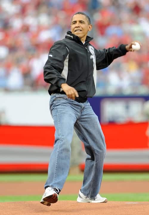 Obama in a zip up jacket, baggy blue jeans, and white sneakers throwing a baseball on a baseball field.