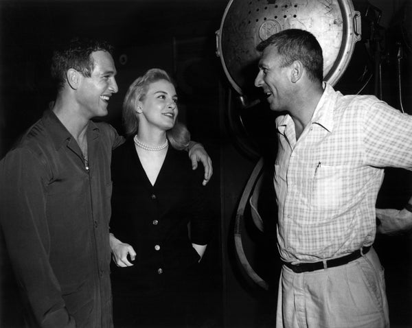 Three people smiling and chatting on a film set, the woman in a classic dress, between two men in casual attire