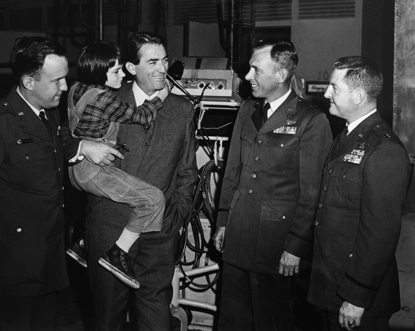 A group of four men in military uniforms and a man in a suit holding a child, engaging in a friendly conversation in an indoor setting