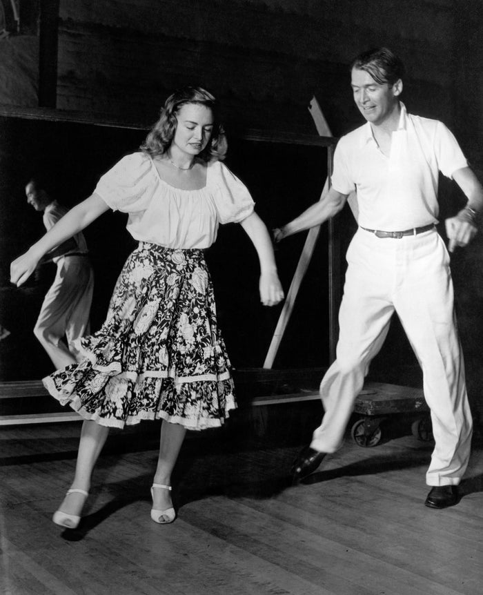 Two people dance in a studio. The woman wears a patterned skirt and blouse, and the man wears a polo shirt and trousers. They appear joyful