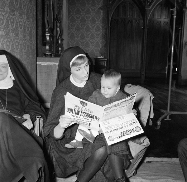 A woman dressed as a nun reads a newspaper with a young child sitting on her lap in a room with another nun nearby