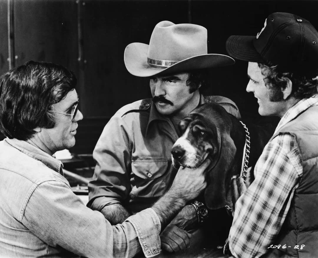 Three men, one in a cowboy hat, gather around a Basset Hound dog. One man gently holds the dog's face as they all look at it, appearing to have a discussion. The image is in black and white.