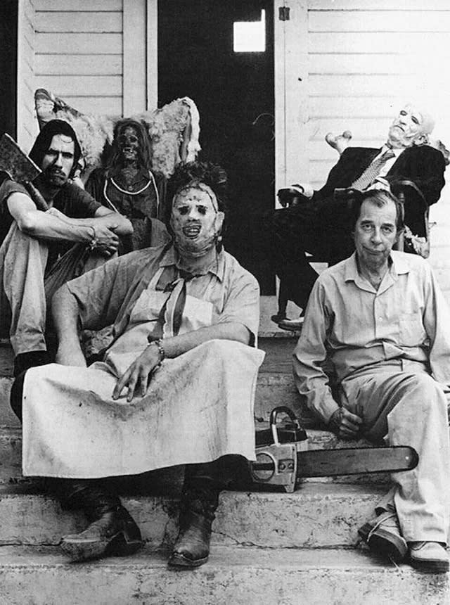 Four people in grim costumes sit on steps in front of a house, one in a leather mask holding a chainsaw, another with a cleaver, and two dressed as eerie figures, with another person slumped in a chair in the background.