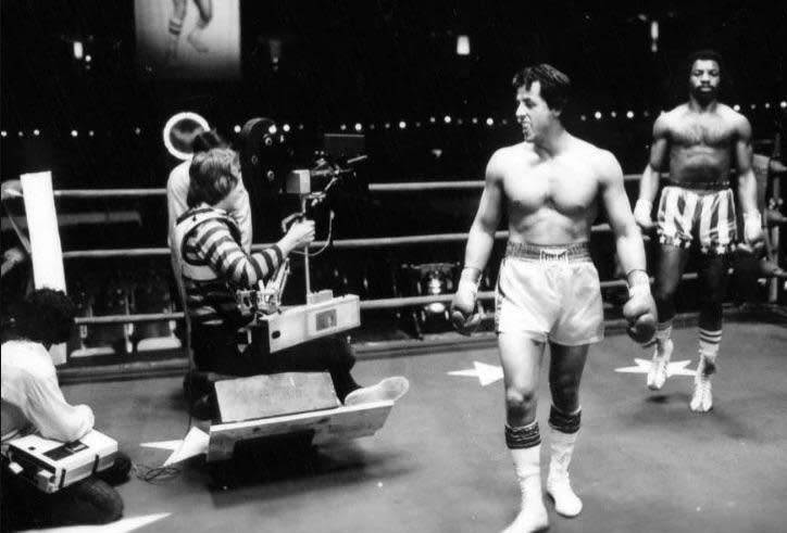 Black and white photo of a boxing ring with two boxers wearing gloves and shorts. A film crew with a camera is capturing the scene, suggesting a movie or TV shoot. The boxers stand ready, with lights and equipment visible.