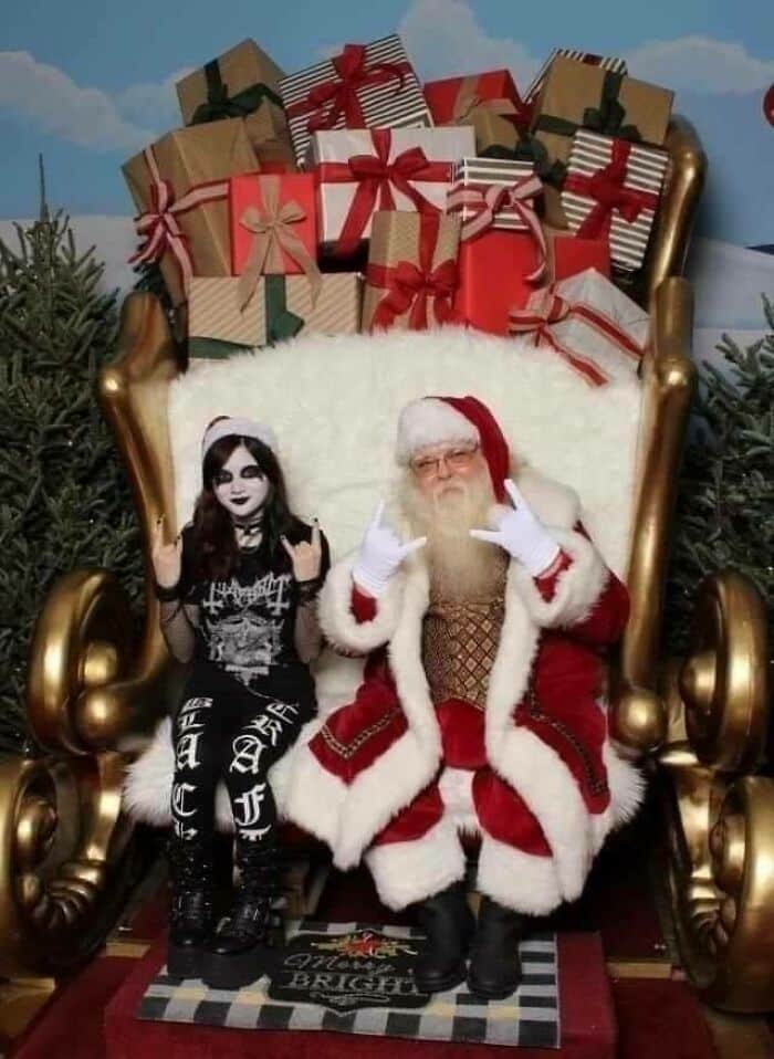 Santa and a person dressed in goth attire make rock hand signs, seated on a chair with gifts, depicting hilarious contrast. Santa and a person dressed in goth attire make rock hand signs, seated on a chair with gifts, depicting hilarious contrast.